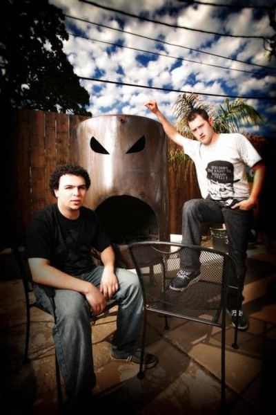 The two members of Dual Boot on a patio, one seated and one standing on a chair, in front of a metal sculpture with angry eyes